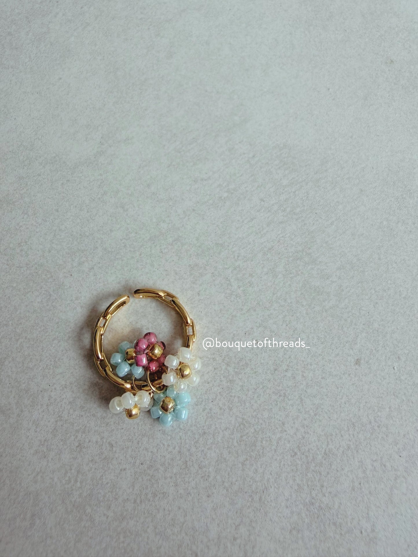 A gold-plated ring featuring a small beaded floral cluster with white, light blue, and pink beads, displayed on a neutral light gray textured background.
