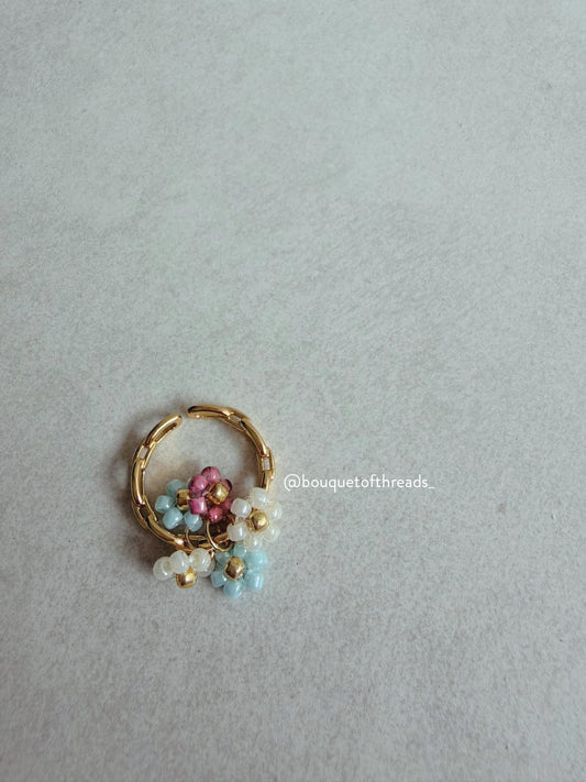 A gold-plated ring featuring a small beaded floral cluster with white, light blue, and pink beads, displayed on a neutral light gray textured background.